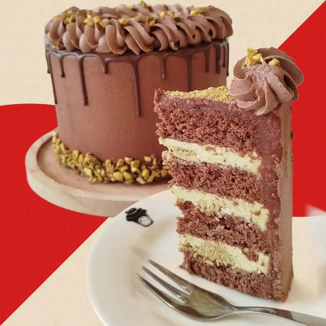 A slice of Dubai Chocolate Pistachio Drip Cake with layers of rich chocolate sponge and creamy pistachio filling, shown in front of the whole cake covered in chocolate buttercream, ganache drip, and sprinkled pistachios.