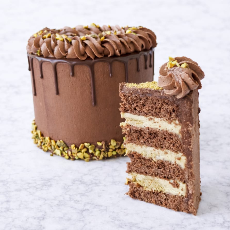 A slice of Dubai Chocolate Pistachio Drip Cake with layers of rich chocolate sponge and creamy pistachio filling, shown in front of the whole cake covered in chocolate buttercream, ganache drip, and sprinkled pistachios.
